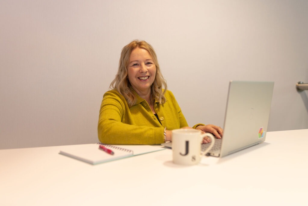 Smiling woman in mustard cardigan working on a laptop, with notebook and J mug on a white desk.