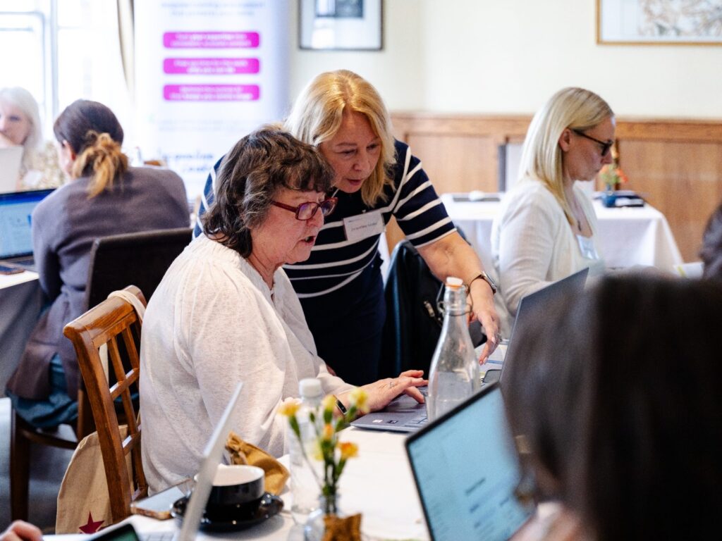 Jacqueline providing one-to-one support to an attendee using a laptop during a group training workshop, with other participants working on laptops in the background.