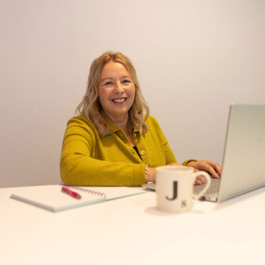 Smiling woman works at a desk with a laptop, notebook and coffee mug in a clean, modern office setting.